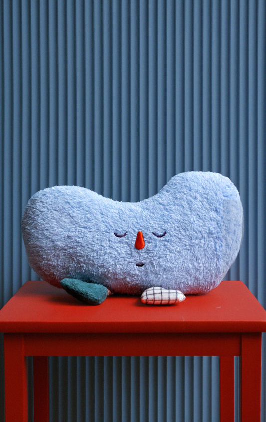 Fuzzy blue pillow with a red nose and green feet on a red table against a blue striped background