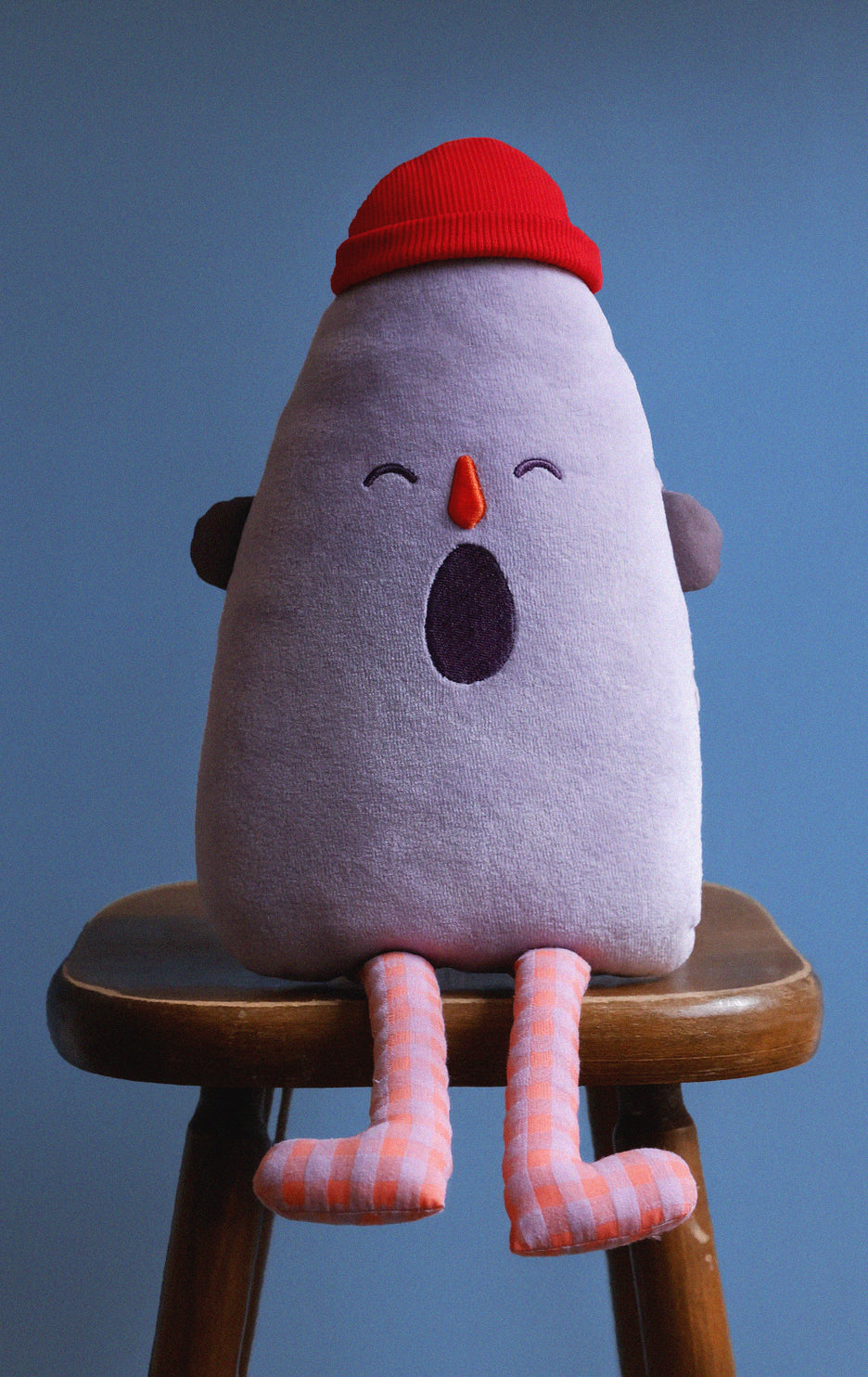 Plush toy with a red hat and striped legs on a wooden stool against a blue background